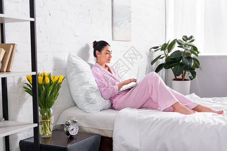睡衣中妇女用膝上型电脑在家中床上图片