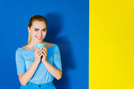 微笑的女孩拿着蓝色和黄色背景上的蓝色杯子图片