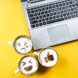 在黄色背景的笔记本电脑旁边的卡布奇诺杯子上涂着微笑的emoji情感和图片