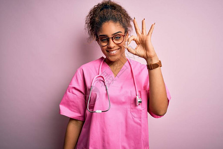 身穿医疗制服和听诊器的非裔美国护士女孩在粉红背景上微笑地用手和指做一个好标记背景图片