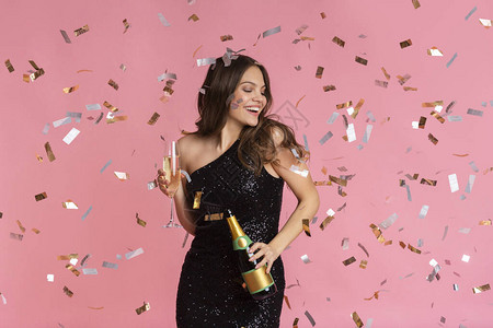 穿着晚礼服的快乐女人拿着香槟酒瓶和装满玻璃的杯子披着粉红背景图片