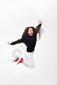 穿着黑帽衫白裤子和红色运动鞋的年轻漂亮的棕发女孩在白色背景中孤立地跳跃快乐的积极背景图片