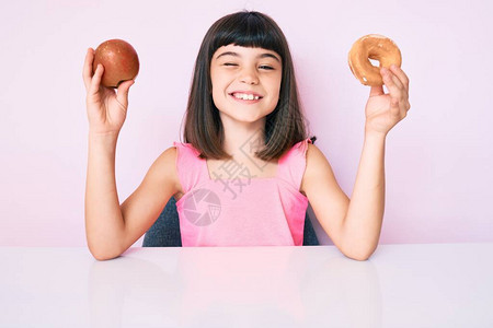 拿着红苹果和甜圈坐在桌上的爆炸女孩用感的表情欢乐和快乐的脸盯着摄影机图片