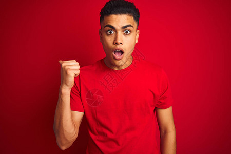 年轻的巴西男子穿着T恤站在孤立的红色背景上惊讶地用手指着一边图片