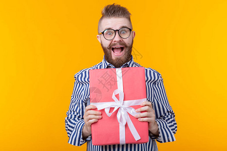 戴着眼镜的小胡子快乐的年轻嬉皮士拥抱黄色背景上的红色礼盒礼物喜悦的概图片