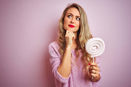 年轻美女在粉红孤立的背景上吃甜糖果面对严肃的面孔思考问题图片