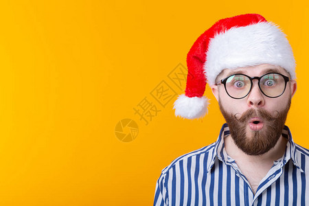 惊讶的年轻人圣诞老人一边看着黄色背景上的相机图片