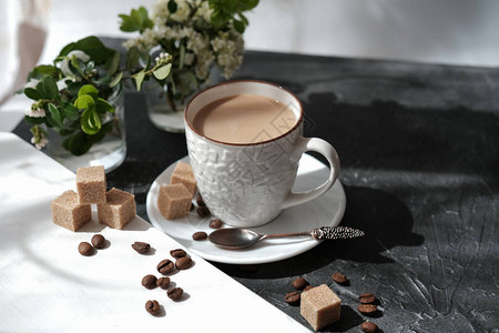 茶杯中的白咖啡棕色糖块和桌子上的咖啡豆花朵和晨图片