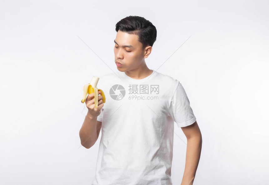 年轻穿白色T恤的亚洲人吃香蕉时面带快乐的脸蛋孤图片