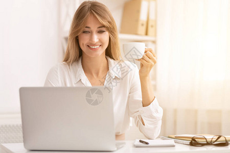 商业生活方式微笑的办公室女孩喝咖啡坐在笔记本电脑在图片