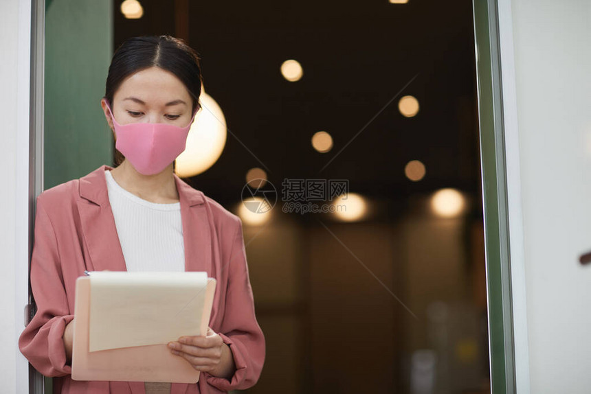 身戴保护面罩的亚洲青年妇女站立并在流行病期间工作的文件上作笔图片