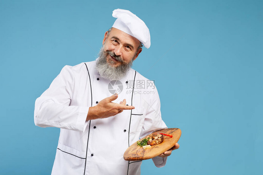 欢快的高级厨师在蓝色背景下站立并在镜头前微笑时在木盘上展示精美菜图片