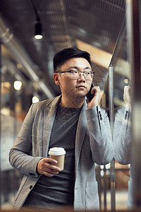 站在现代办公室内玻璃墙壁上站立的亚洲青年商人通过电话发言图片
