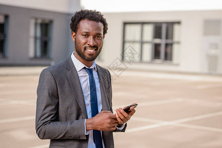 英俊的非洲美籍商人持有智能手机看相机和笑图片