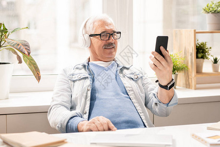 当代穿便装和耳机的成年流动男子在办公期间观看智能手机视频图片