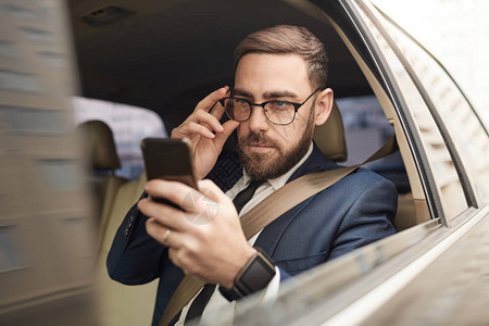 戴眼镜的年轻商人坐在汽车后座上看着手机屏幕图片