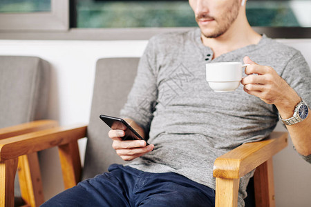 男人坐在扶手椅喝茶杯和阅读智能手机新图片
