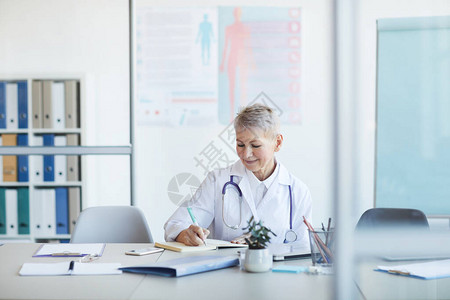成熟女医生写笔记和微笑时坐在室内诊所复印室的桌子图片