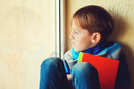 悲伤的男孩坐在窗罩上孩子望着窗外有书的哀伤的男小学生靠近窗户的体贴的孩子独自伤心的孩子家暴负面情绪抑郁背景图片