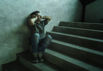 年轻抑郁和偏执的男人坐在户外黑暗的垃圾街角楼梯上的戏剧肖像图片