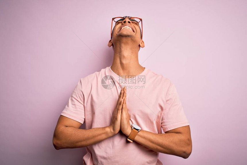 身穿轻便T恤和眼镜的英俊的非洲男子在粉红色背景下乞讨亲手祷告以及脸上充满希望的表情图片