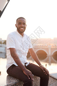 身穿白衬衫坐在大理石栏杆上靠着城市河流微笑的留着胡须的非洲裔美国背景图片