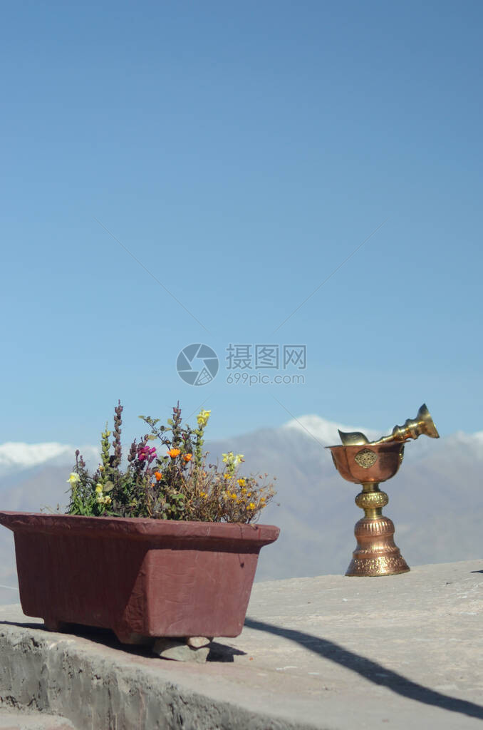 两座金铜礼仪杯放在一座佛教寺庙的石架上他们旁边是一个装满鲜花的红色花盆远处是西喜马拉雅山脉的白雪皑的山图片