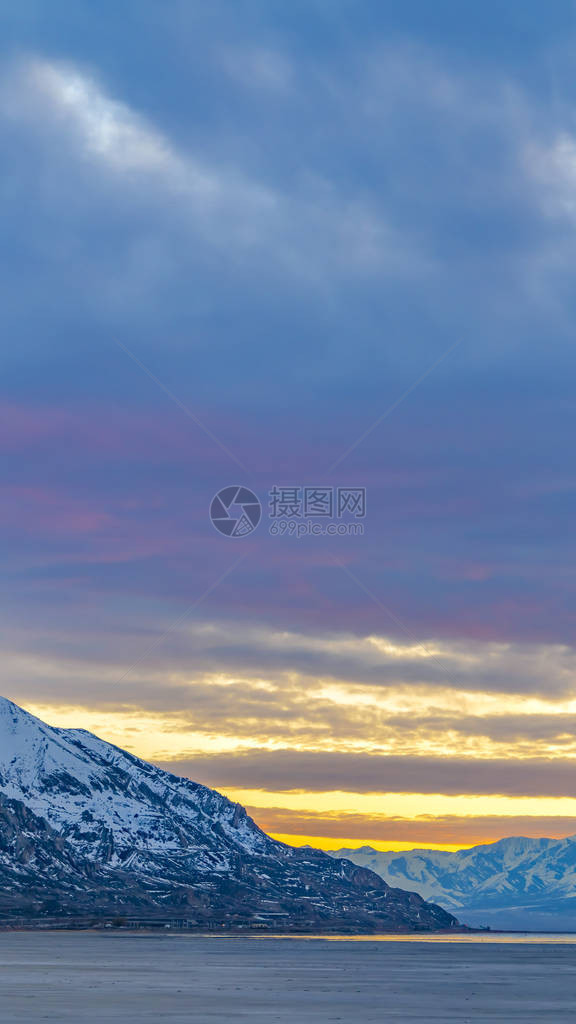 平静的湖面和被雪覆盖的雄伟山脉的垂直框架全景头顶乌云密布的天空在日落时被金图片