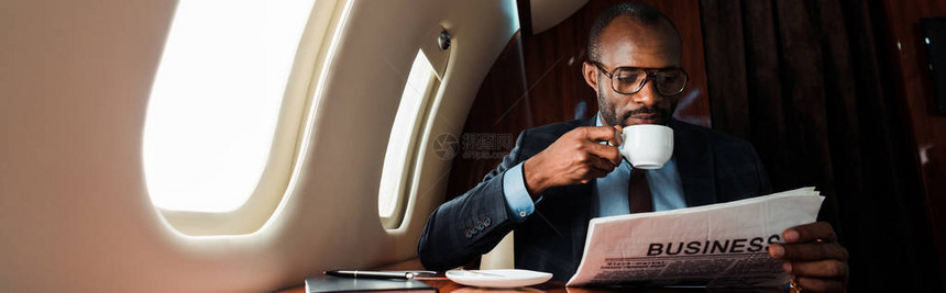 非洲裔美国商人戴着眼镜在私人飞机上拿着杯子阅读商业报图片