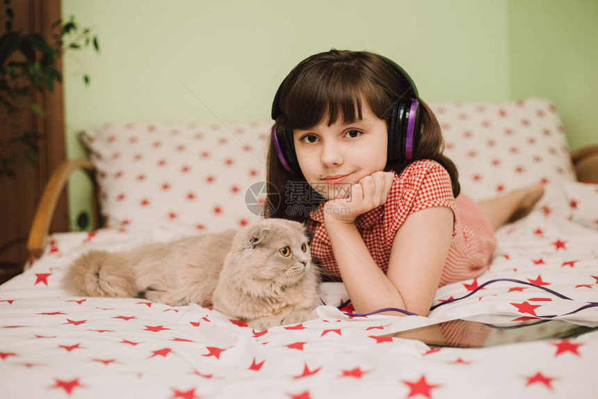 戴着耳机听音乐的女孩和漂亮的毛茸的猫躺在女孩附近使用平板电脑的孩子在冠状期间图片