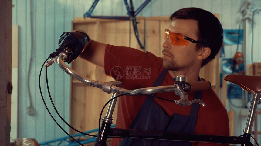 主题小型企业自行车维修一位戴着安全眼镜手套和围裙的年轻白种人黑发男子坚持在车间库的自行车把上图片