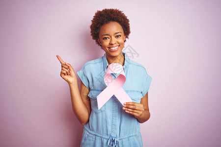 年轻非洲裔美国女在孤立的粉红背景上握着浅色癌症丝带快乐地用手和图片