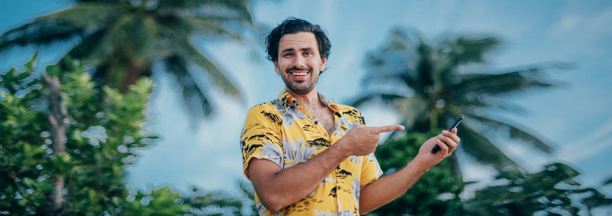 一个男人手里拿着电话站在棕榈树的背景上一个年轻人用手指着智能手机他是热带岛屿上的夏威夷衬衫英俊的男人和明图片