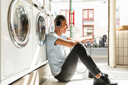 快乐的北非男子坐在地板上靠在洗衣店的洗衣机上用手机和耳机听音图片