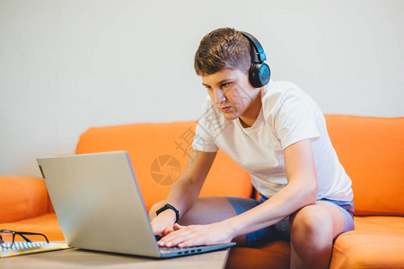 穿着白色T恤的可爱少年坐在笔记本电脑和书房旁边的橙色沙发上戴耳机的严肃男孩做作业背景图片