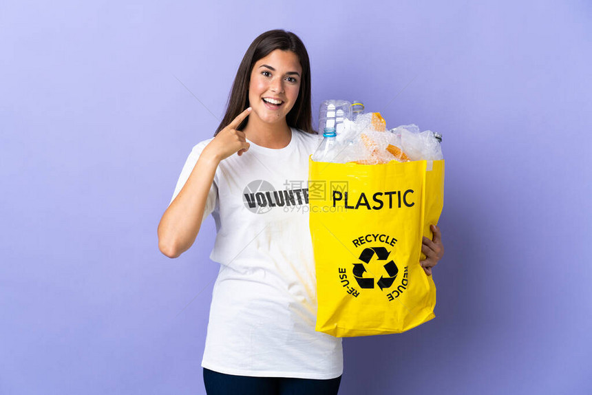 拿着一袋装满塑料瓶子的年轻巴西女人用紫色背景隔离的玻璃瓶回收利用图片