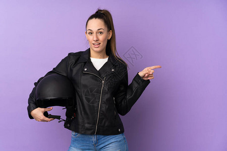 带着摩托车头盔的年轻黑发女人与孤立的紫色背景相隔绝惊讶并用手图片