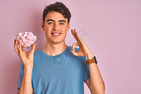 十几岁的男孩在孤立的粉红色背景上拿着一堆美味的蓬松棉花糖图片