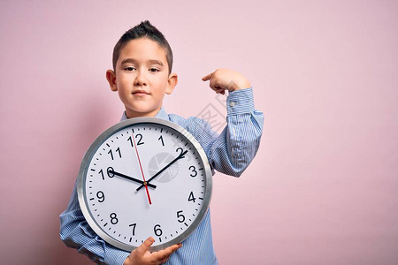 年轻小男孩在孤立的粉红背景中抓着一个大钟头拿着巨大的时钟并用奇异图片