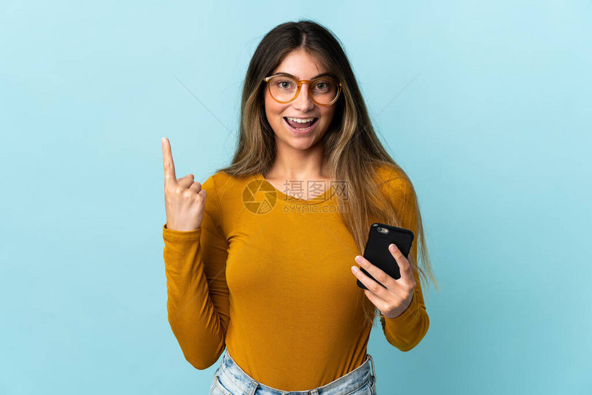 使用蓝色背景中孤立的手机的年轻白人女打算在举起手指的同时图片