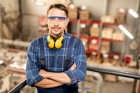 自信的年轻跨武装工程师在现代大型工厂里通过保护眼镜看着图片