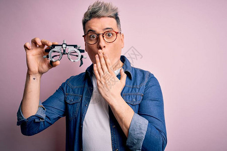 年轻的现代光学验光师男子拿着验光眼镜在粉红色的背景上盖着嘴图片