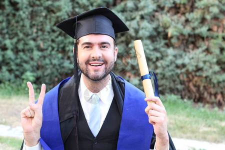 英俊的青年学生穿着毕业服装在公园获得滚版文凭的背景图片