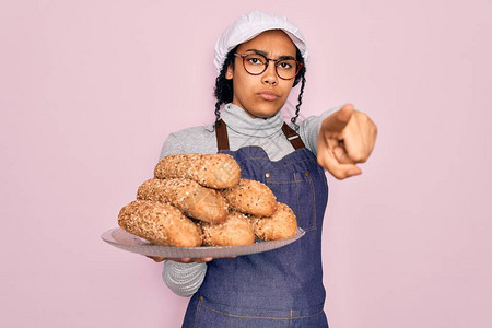 年轻的非洲裔美国面包师妇女穿着围裙拿着全麦面包和麦片用手指着相机和你图片
