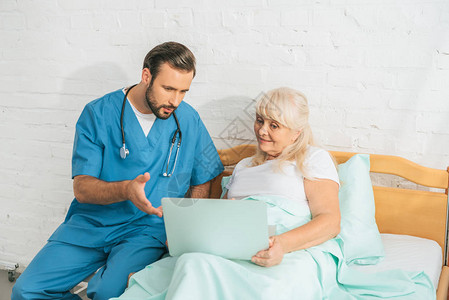 在医院床上使用膝上型看老年妇女的男护士有听诊图片