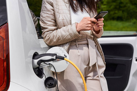 年轻女子手持机站在电动车旁等待汽车电池充电图片