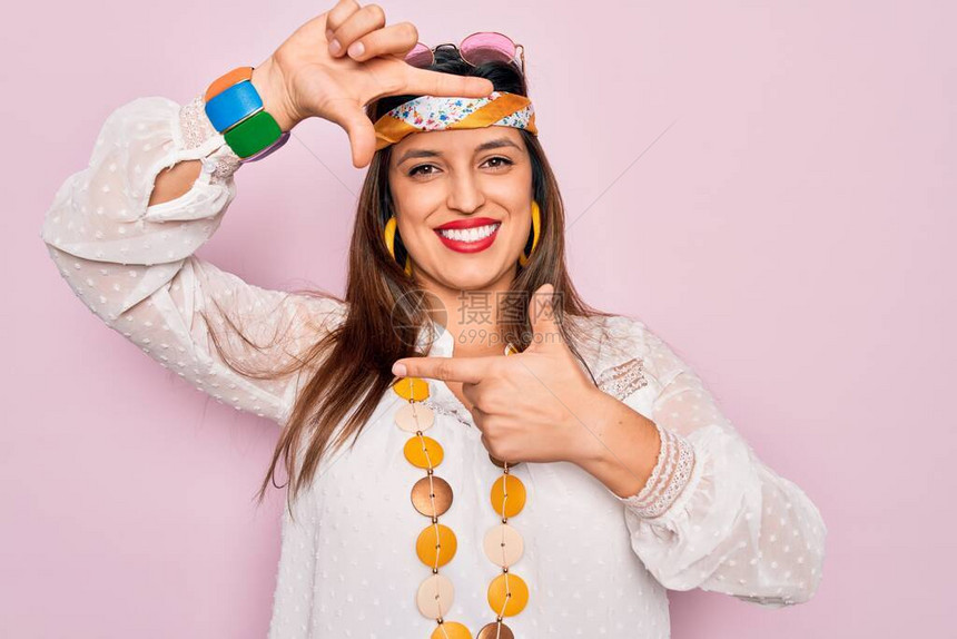 穿着时装布霍风格和太阳镜的年轻西班牙籍嬉皮女青年在粉红色背景上微笑地用手和指搭架图片