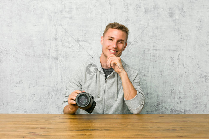 年轻摄影师拿着摄像头在桌子上微笑着快乐和自信图片