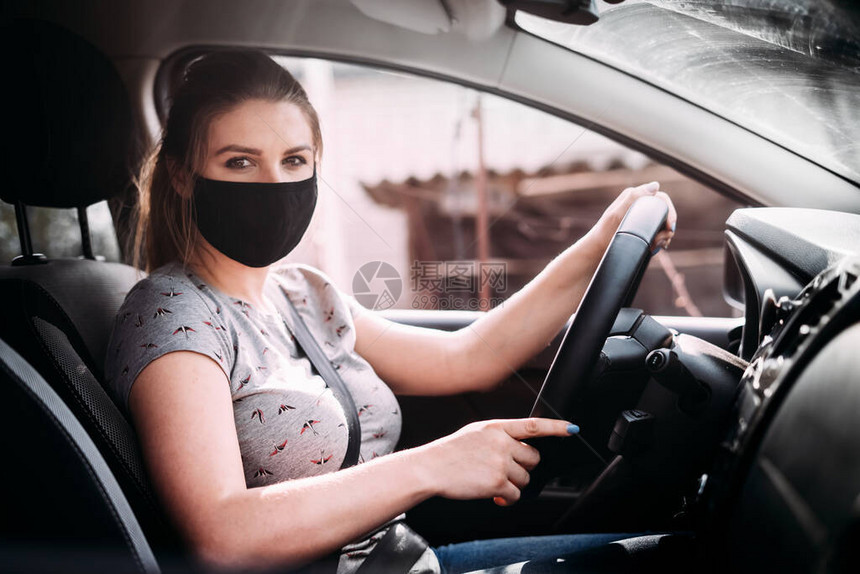 一名戴着黑色医用口罩身穿灰色T恤金发的年轻女子坐在左驾汽车上她的手放在方向盘上看着相机人图片