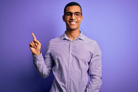 身穿条纹衬衫和眼镜面带大笑脸的紫色背景的非洲帅哥美国人用手指向镜头的侧面背景图片